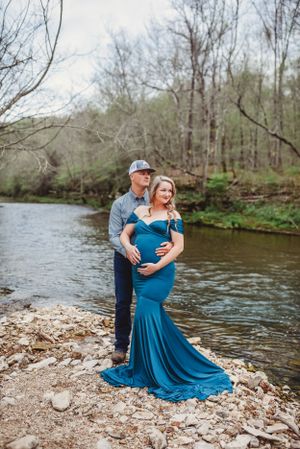 Couple in blue dress and casual wear pose romantically by riverside on rocky shore.