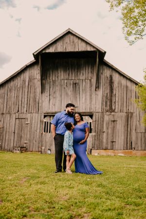 A maternity photoshoot in front of a weathered wooden barn on a grassy hill.