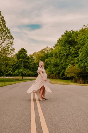 A couple in light colored outfits walks along a winding road surrounded by green trees on a cloudy day.