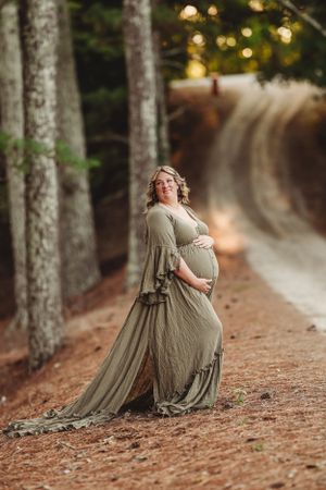 A sage green maternity dress photoshoot in a wooded path with sunlight filtering through trees.