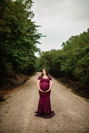 A woman in a burgundy dress poses gracefully on a winding gravel path through the woods.