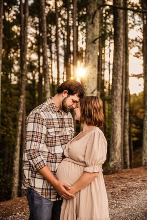 Romantic maternity photo in forest with golden sunlight filtering through tall pine trees.