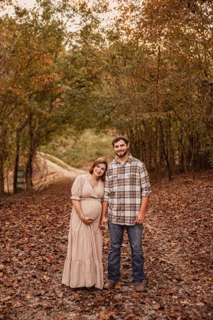 Autumn maternity photo on leaf-covered path with rustic woodland backdrop.
