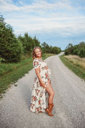 Maternity portrait on a country road wearing a white floral dress with brown boots.