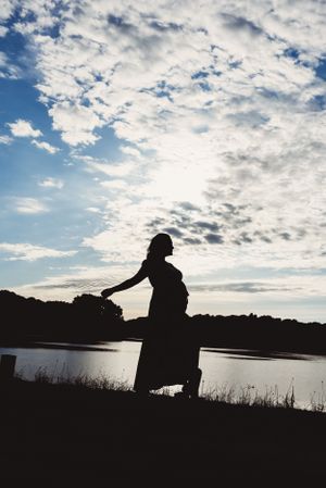 Silhouette of an expectant mother against a dramatic cloudy sky by a lake at sunset.