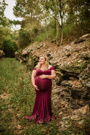 Expectant mother in burgundy gown poses elegantly against rocky hillside in natural setting.