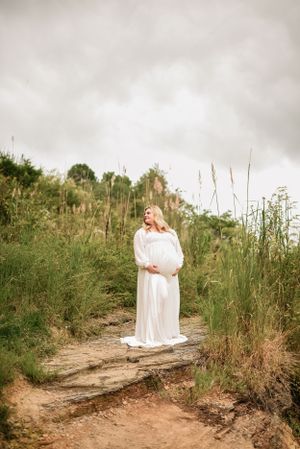 Ethereal maternity portrait of woman in flowing white dress on nature path.