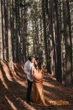A couple shares tender moments in a pine forest with dramatic sunlight filtering through the trees.