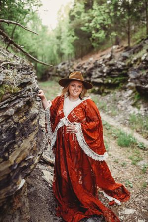 Figure in red vintage dress and brown hat poses among forest rocks.