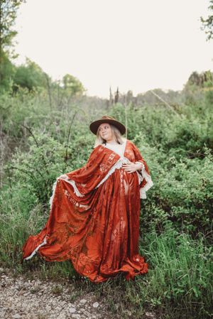 Person in a flowing red vintage dress posing dramatically in a green field.
