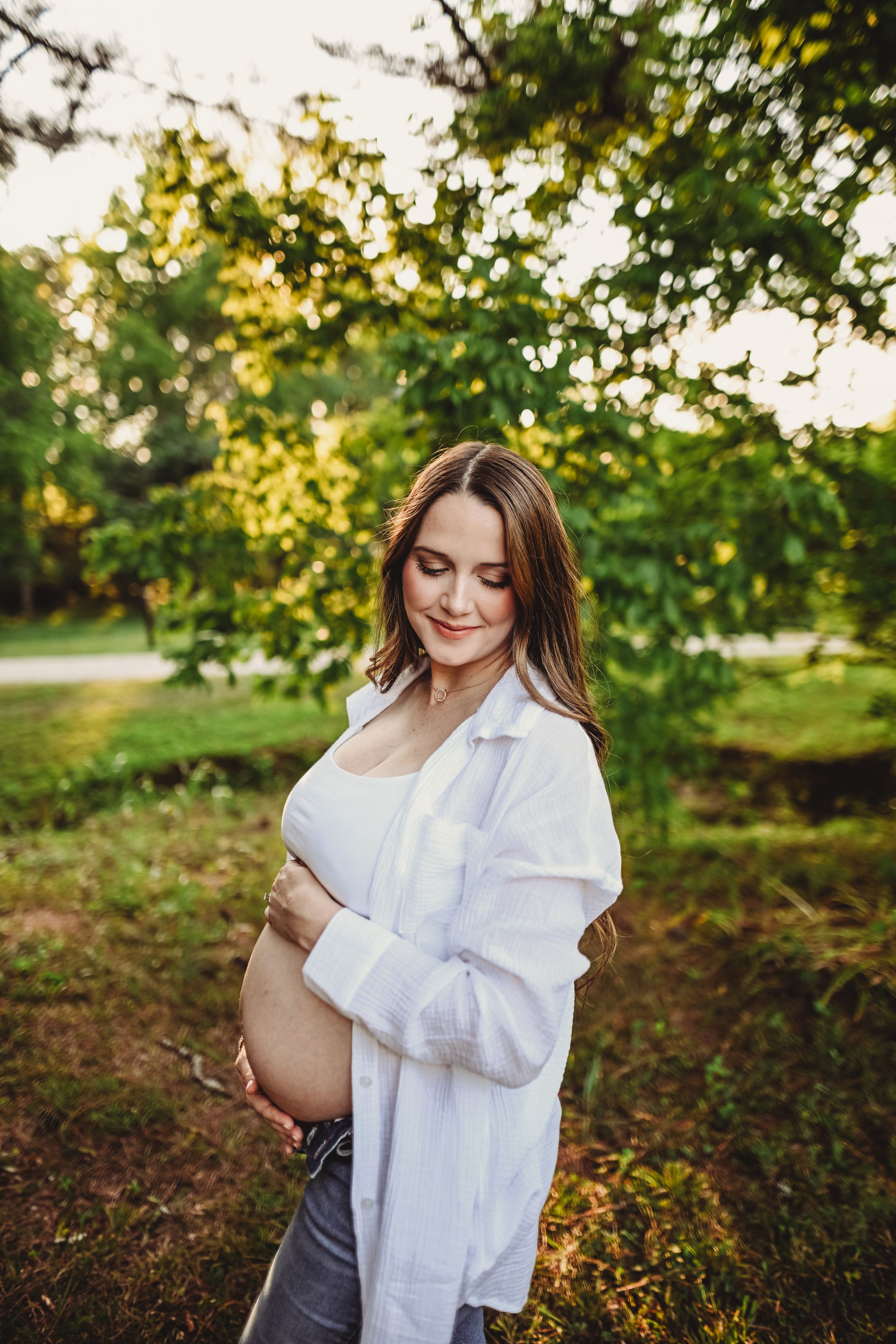A maternity portrait in a white cardigan against a sunlit garden backdrop.