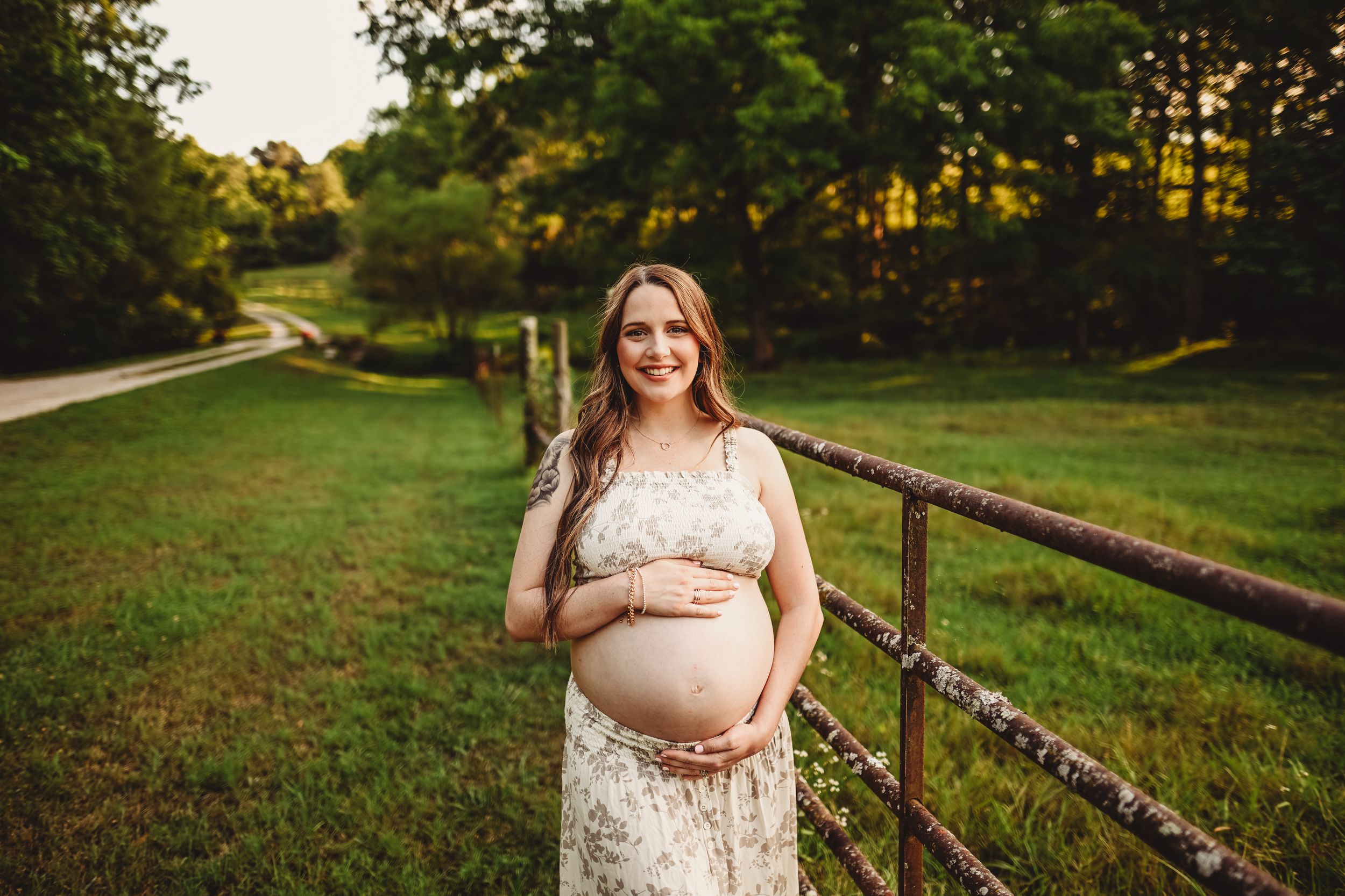 A maternity portrait in a lace dress against a rustic wooden fence at sunset.