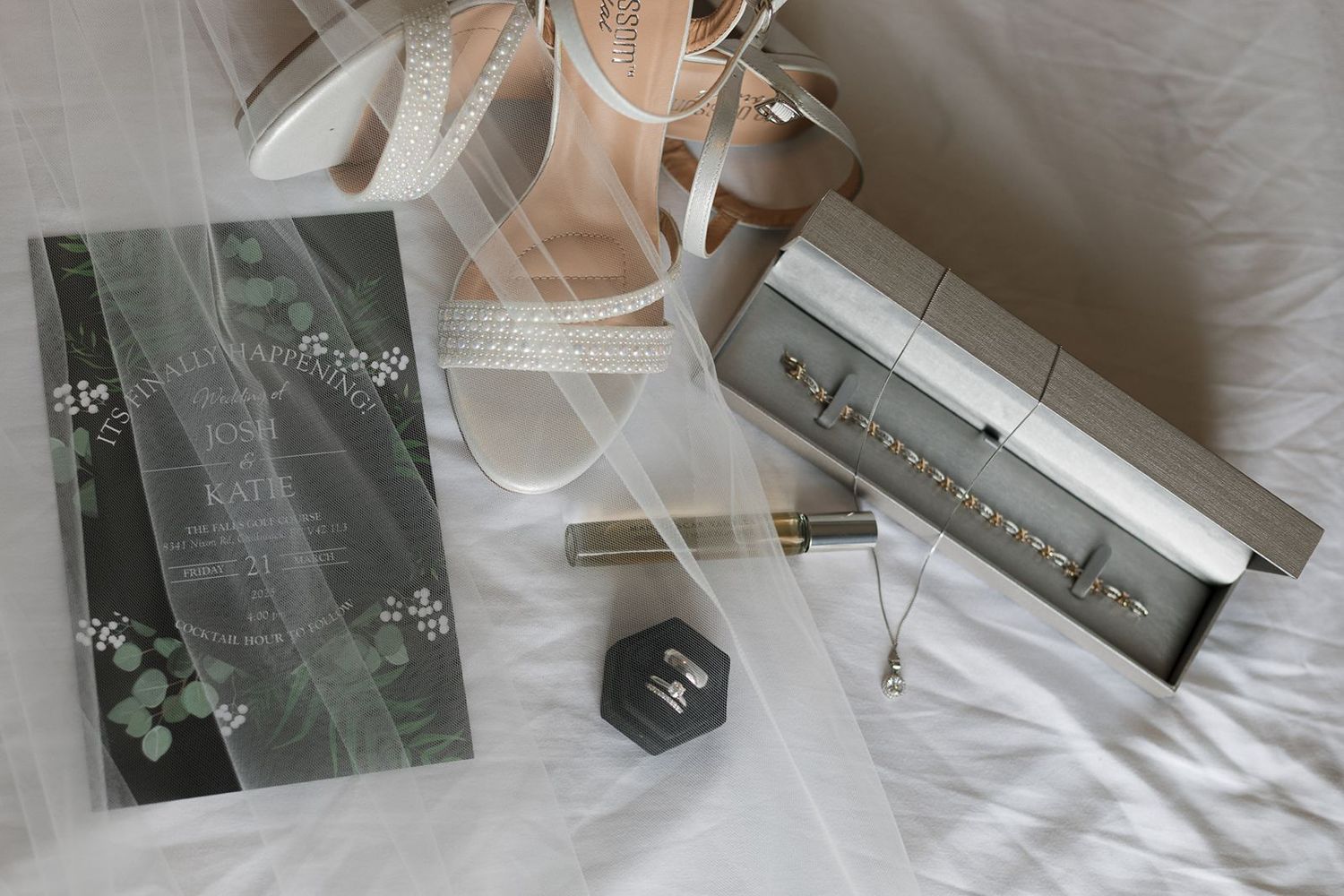Wedding flat lay featuring bridal veil, nude heels, jewelry, perfume, and invitation on white fabric.