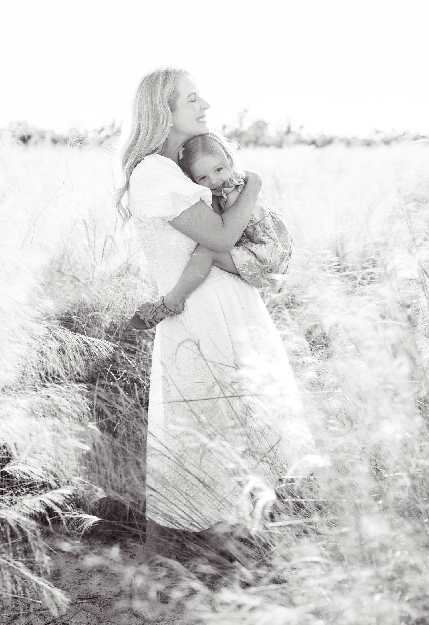 A couple stands together on a grassy hill with sunlit trees in the background during a maternity photo session.