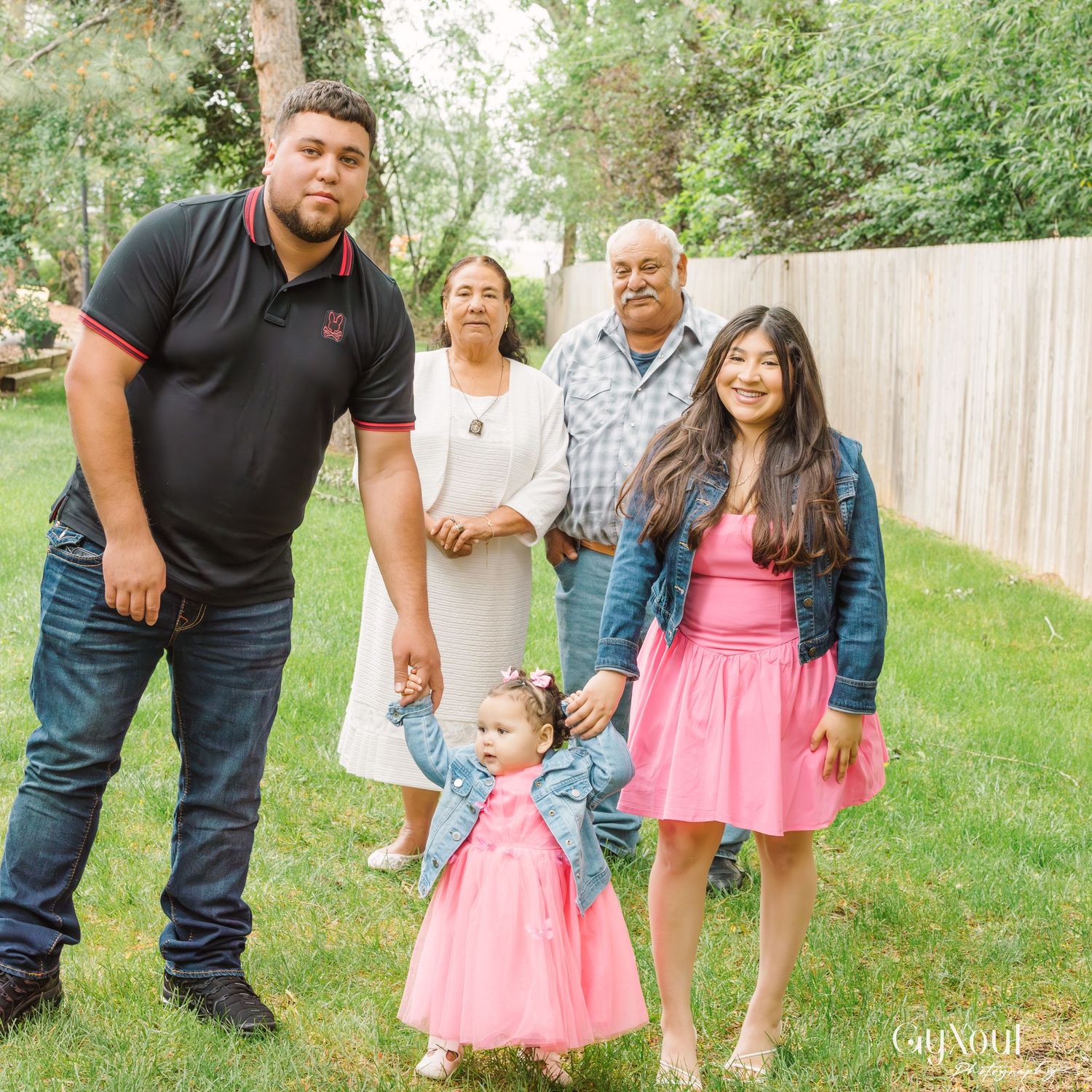 A family gathers outdoors on a grassy lawn near a wooden fence while posing for candid lifestyle photos.