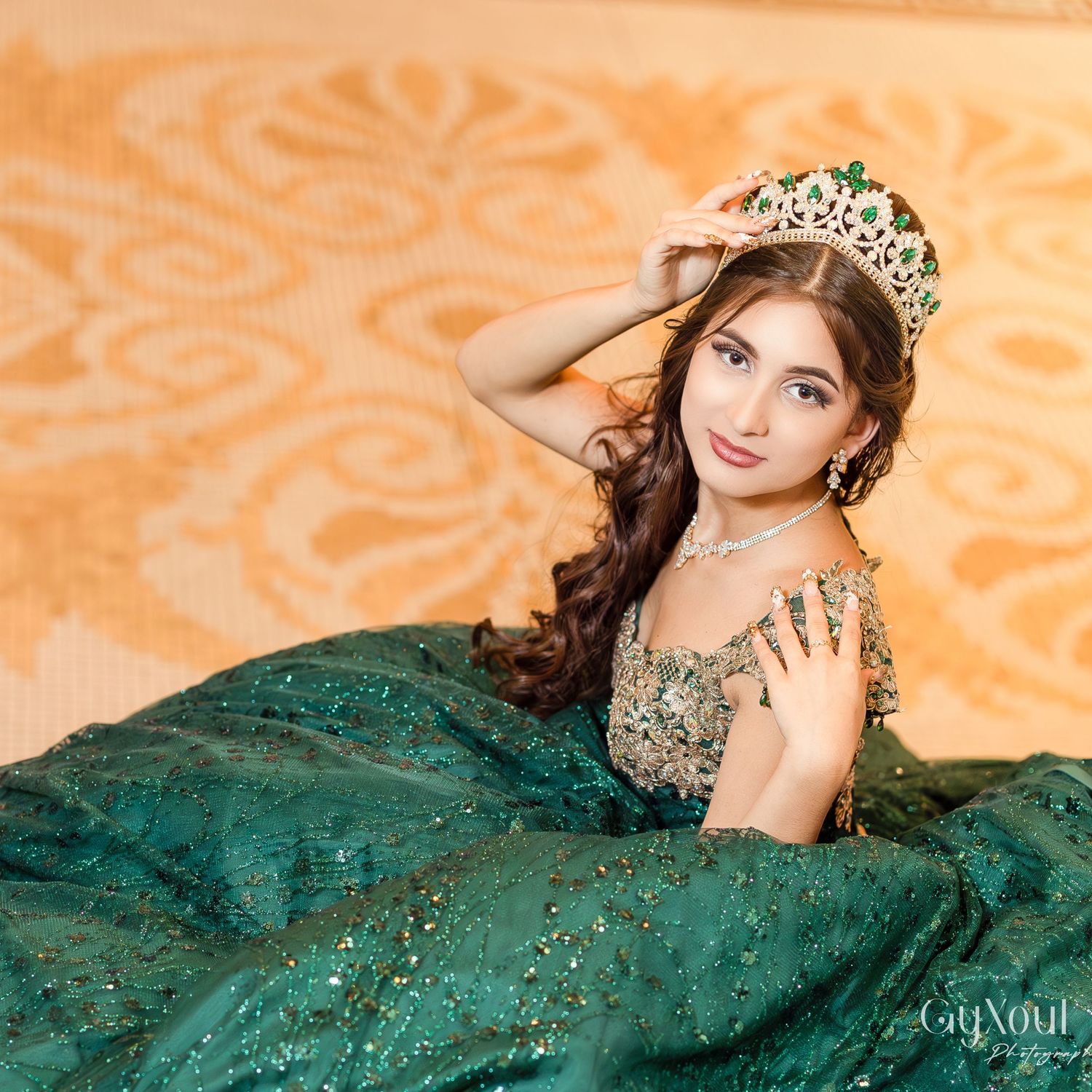 A person in an elegant emerald green quinceañera ball gown with a jeweled crown poses against an orange patterned backdrop.