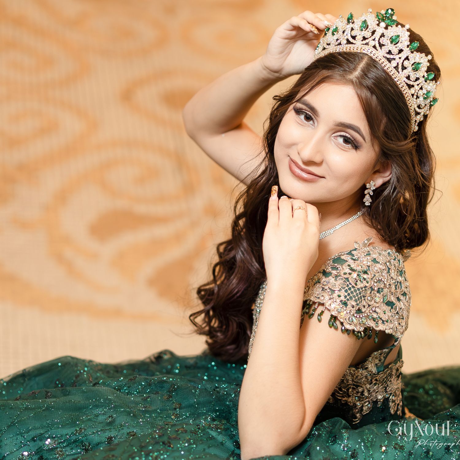 A pageant contestant wearing an ornate crown and emerald green evening gown poses elegantly against a peach background.