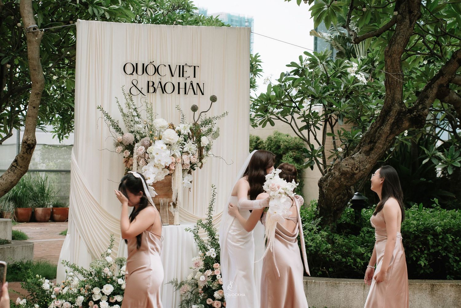 Bridesmaids in blush dresses prepare near a wedding backdrop reading Quoc Viet and Bao Han.