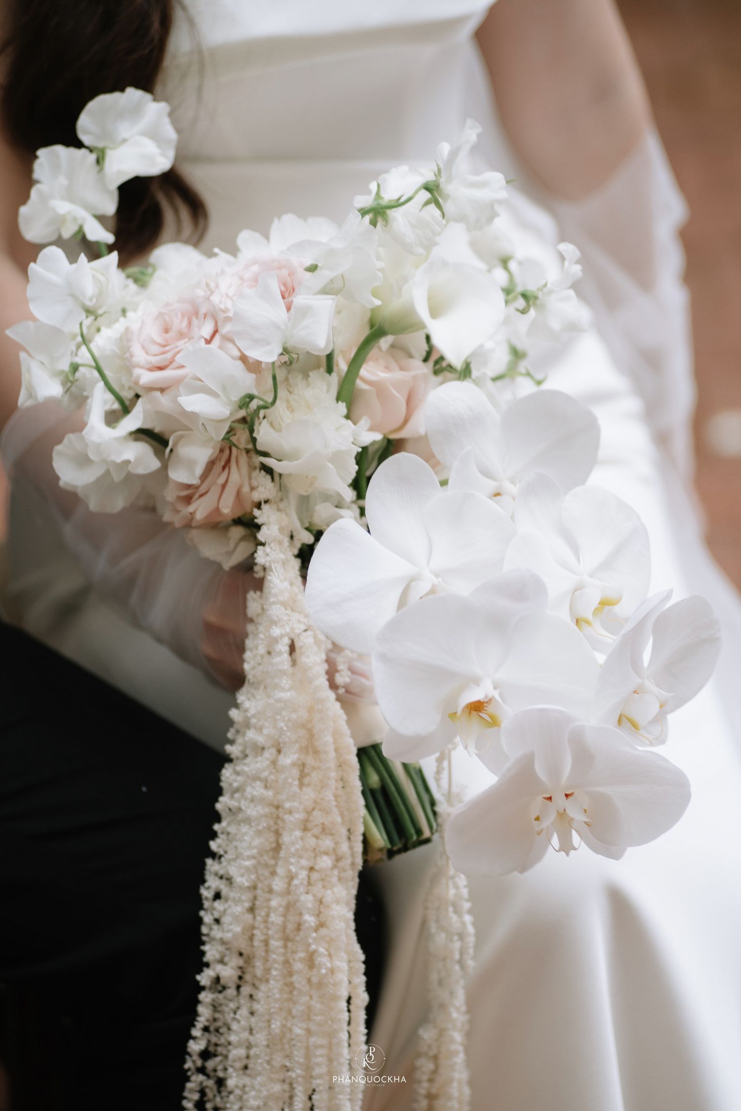 Elegant bridal bouquet featuring white orchids, blush roses, and cascading pearl-like details held by bride in white gown.
