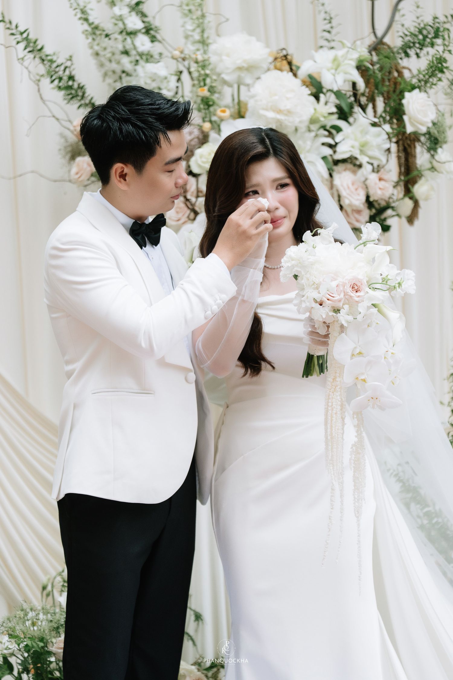 Emotional groom in white tuxedo gently wipes tears from bride's cheek during romantic wedding ceremony.