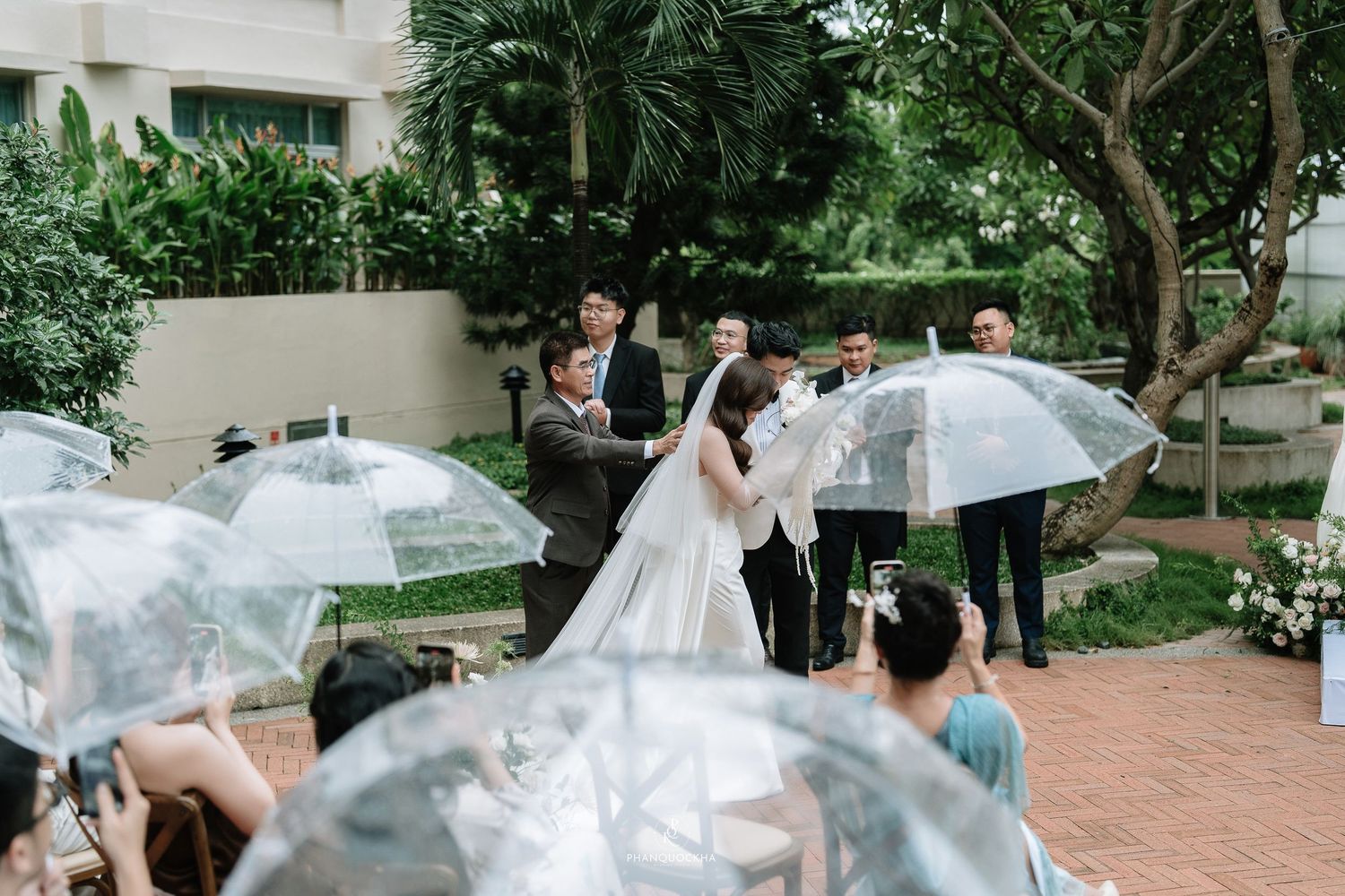 Bride in white gown with long veil stands before guests holding clear umbrellas at outdoor ceremony.