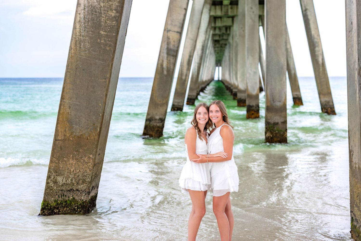 Two people in white dresses pose under a pier while ocean waves roll in on a sunny beach day.
