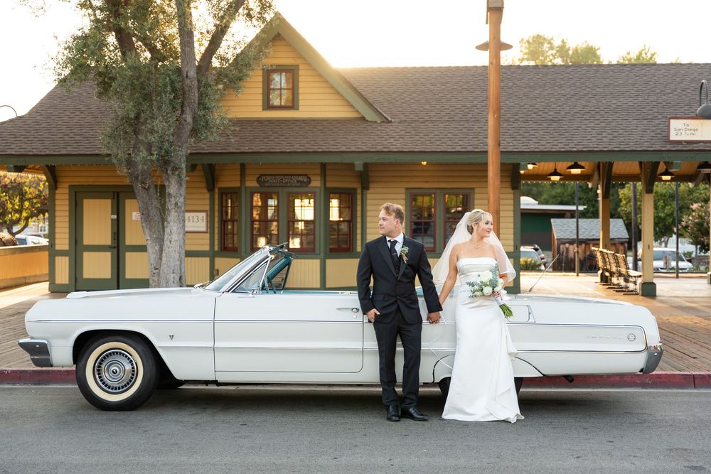 Wedding couple stands by a classic white vintage Lincoln Continental limousine outside a venue.