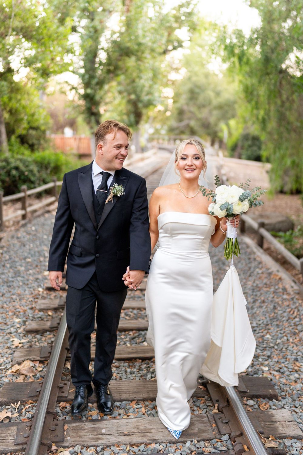 A newlywed couple walks together on rustic train tracks surrounded by lush green trees while holding hands.