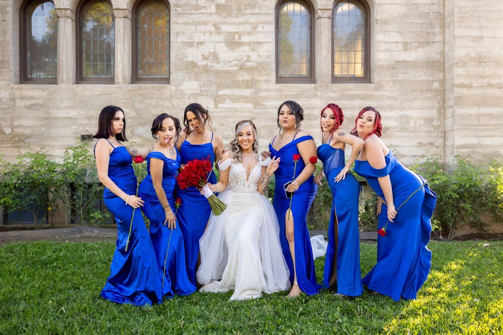 A group in matching royal blue dresses pose with a bride in white on a sunny day outside a stone church.