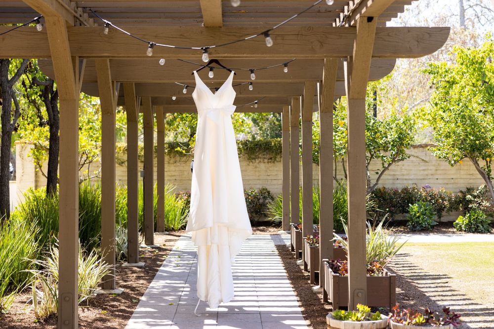 White wedding dress hanging on wooden pergola in garden setting with natural sunlight streaming through.