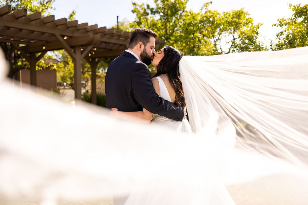 A couple embraces romantically under a pergola with flowing white veil in golden sunlight during their wedding.