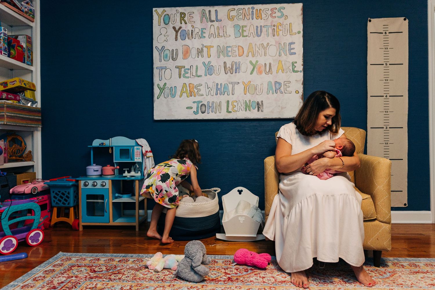 A nurturing quiet moment in a colorful playroom with a child playing in the background and nursing scene in a yellow chair.