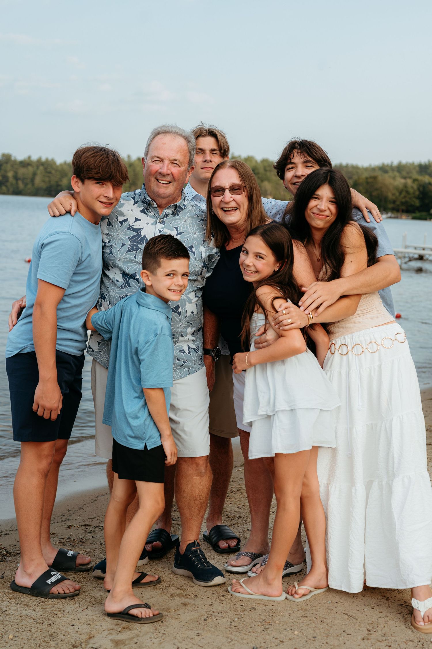 A multigenerational family smiles and embraces together on a sandy lakeside beach.