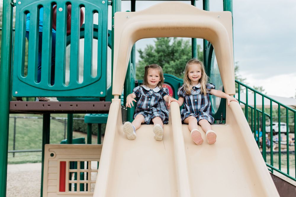 Two children enjoy sliding down parallel tube slides at an outdoor playground with green equipment.