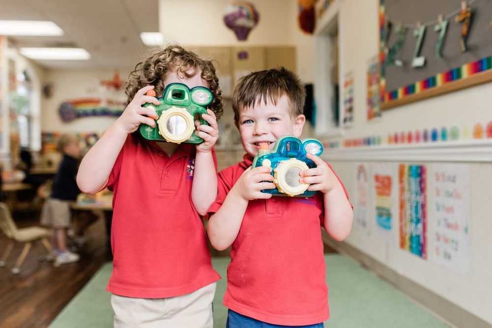 Two children in red shirts taking pictures with toy cameras in a classroom hallway.