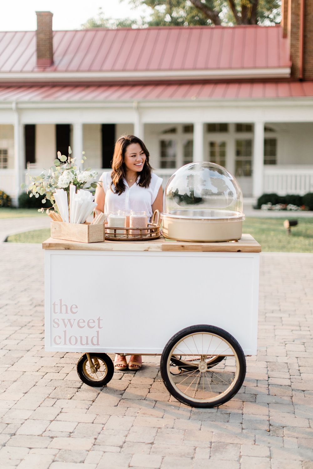 A white dessert cart with 'The Sweet Cloud' branding and glass display domes stands on brick pavers outside a house.