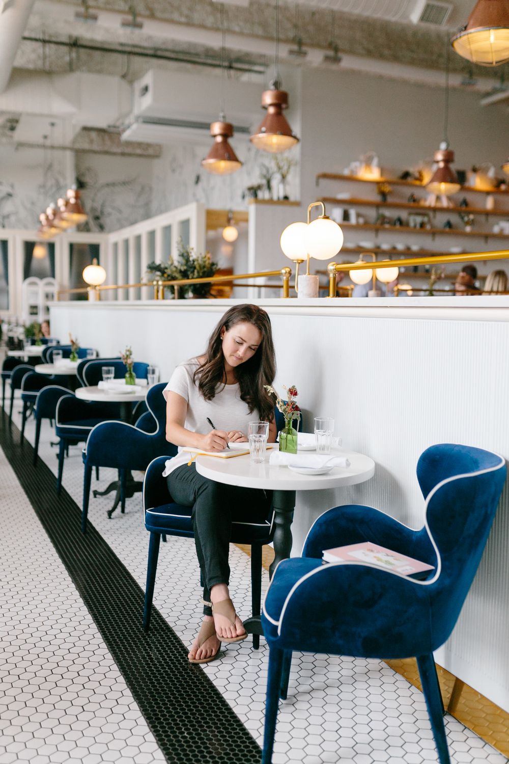 Interior of trendy restaurant with blue velvet chairs, white marble tables and vintage pendant lighting fixtures overhead.