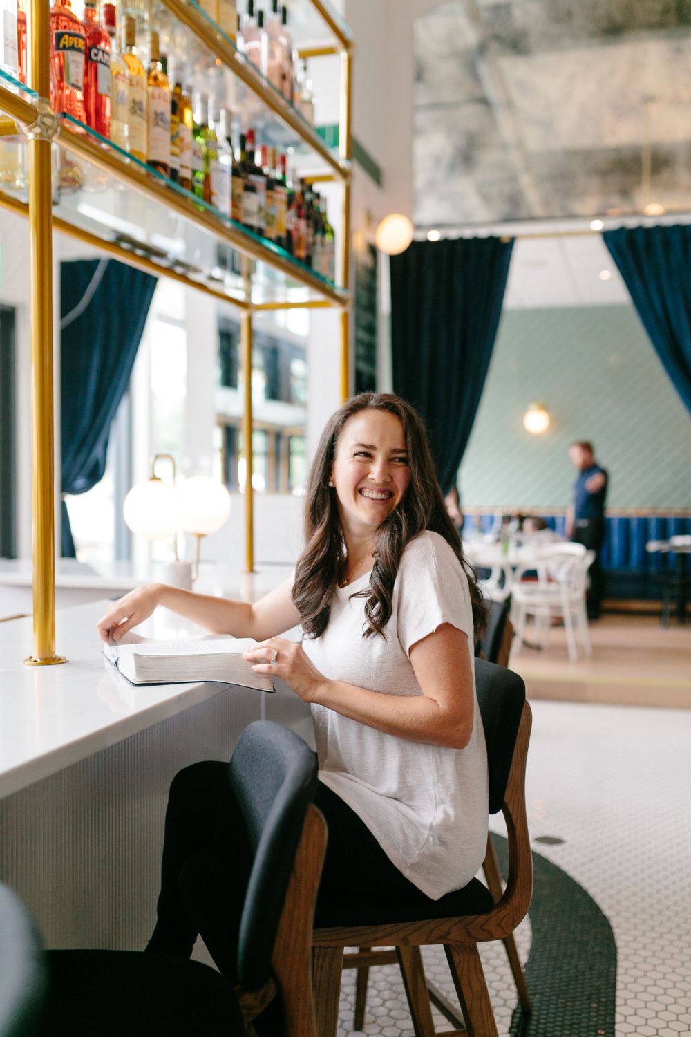 Guest sitting at a bright restaurant bar with shelves of liquor bottles and blue curtains in the background.