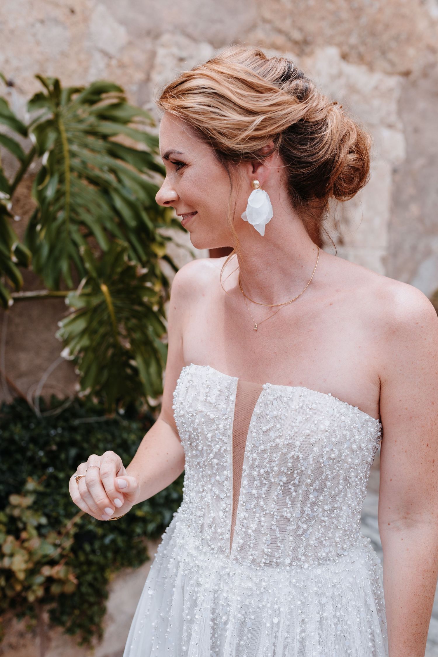 Ein elegantes Brautkleid mit zarten Perlenstickereien wird durch weiße, blattförmige Ohrringe und eine hochgesteckte Frisur ergänzt.