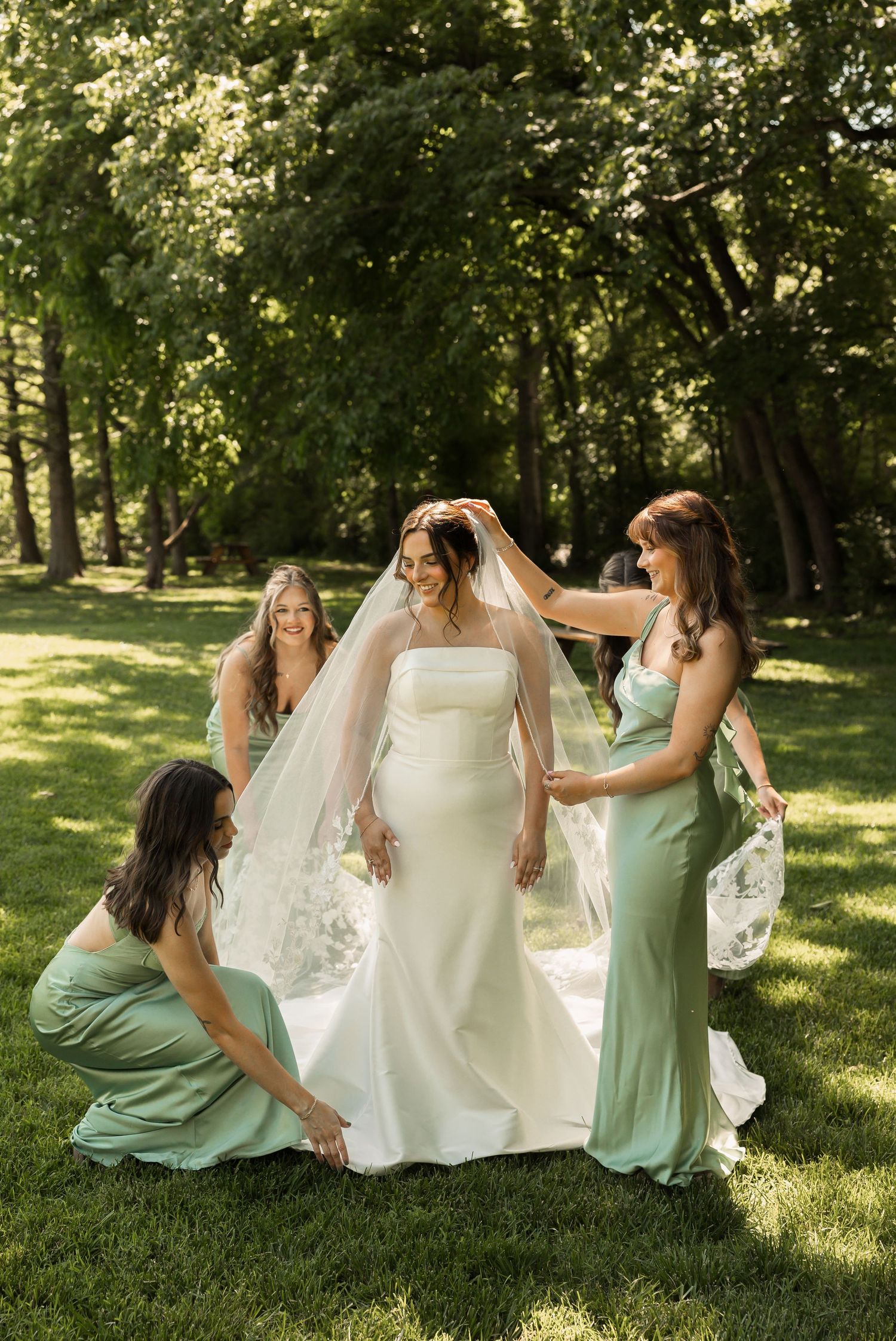Bridesmaids in sage green dresses help adjust the bride's veil and gown during an outdoor summer wedding photoshoot.