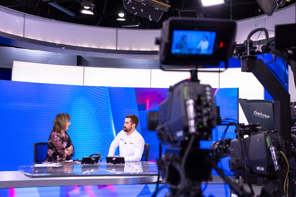 Behind the scenes view of a television broadcast studio with a camera filming two people at a news desk with blue backdrop.