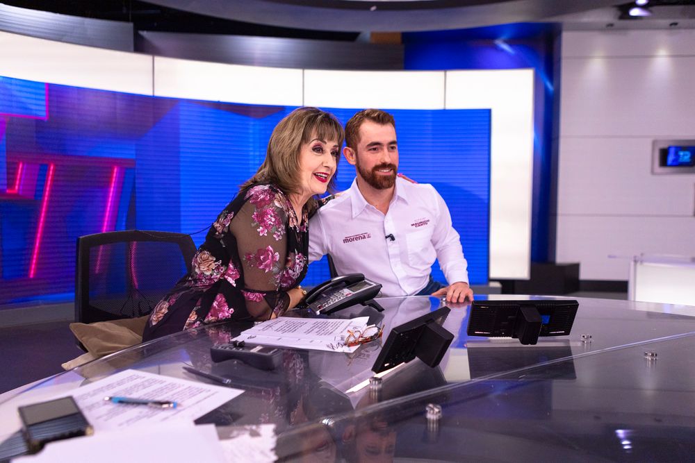 Two television presenters share a lighthearted moment at a news desk with blue studio lighting in the background.
