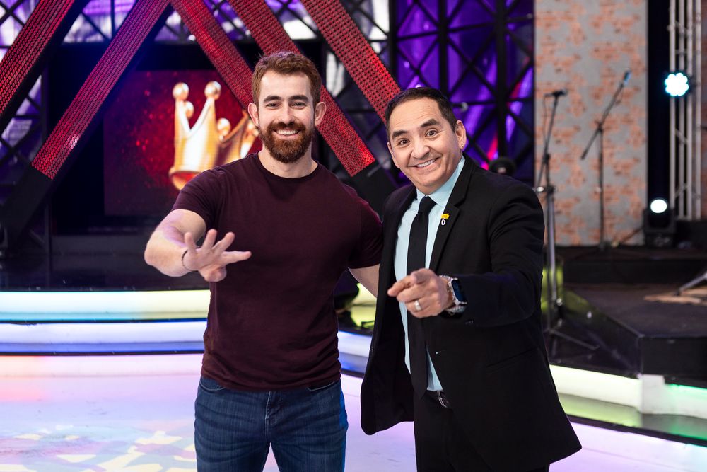 Two people stand together smiling on a brightly lit stage with a purple backdrop and stage lighting visible.