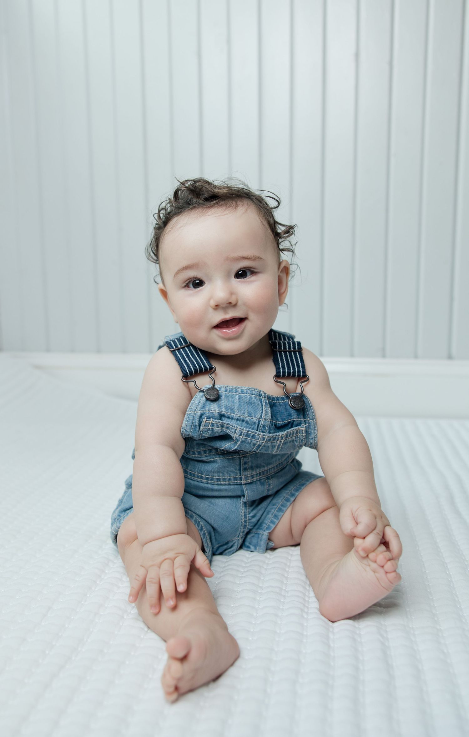 Cheerful baby wearing blue denim overalls sits on white textured surface against light background.