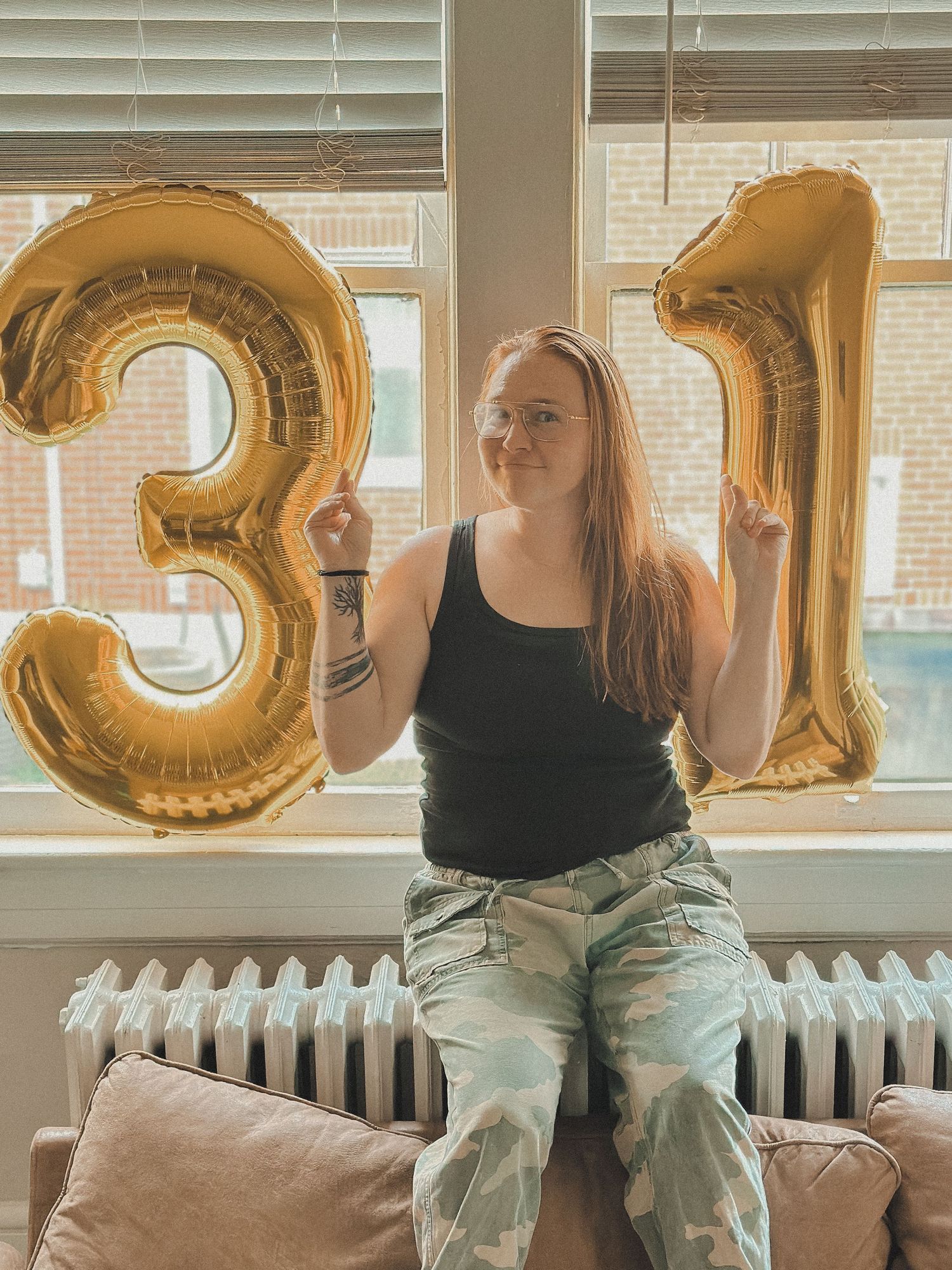 Woman in camo pants and black tank top poses with gold "31" balloons by a window, smiling and pointing upward.