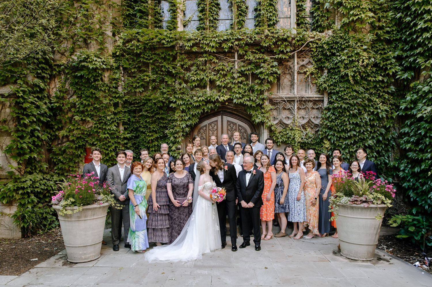 Large group gathered for wedding photo outside ivy-covered stone building with elegant potted plants framing the entrance.