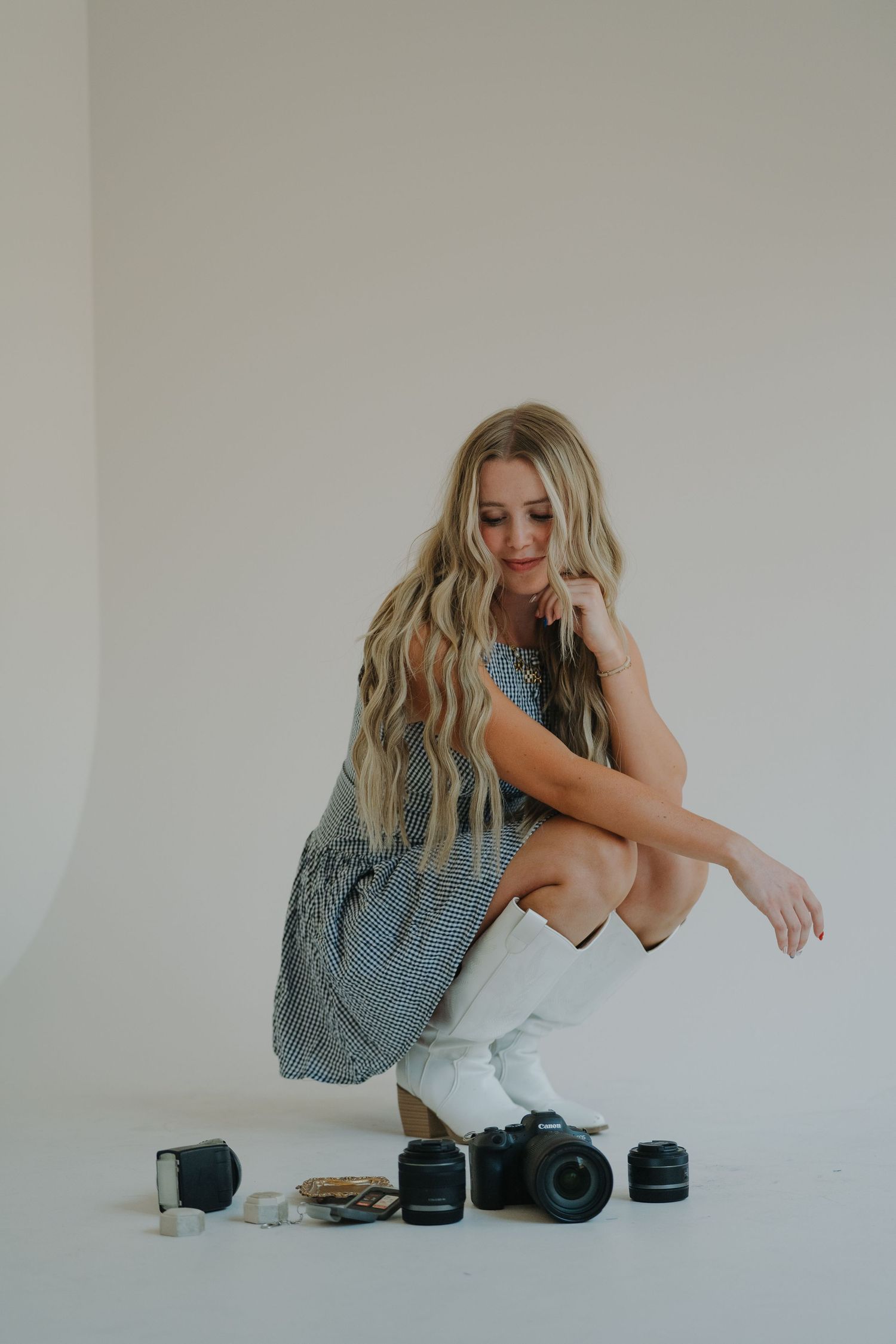 Blonde woman crouching beside Canon camera, lenses, and photography gear on white background.