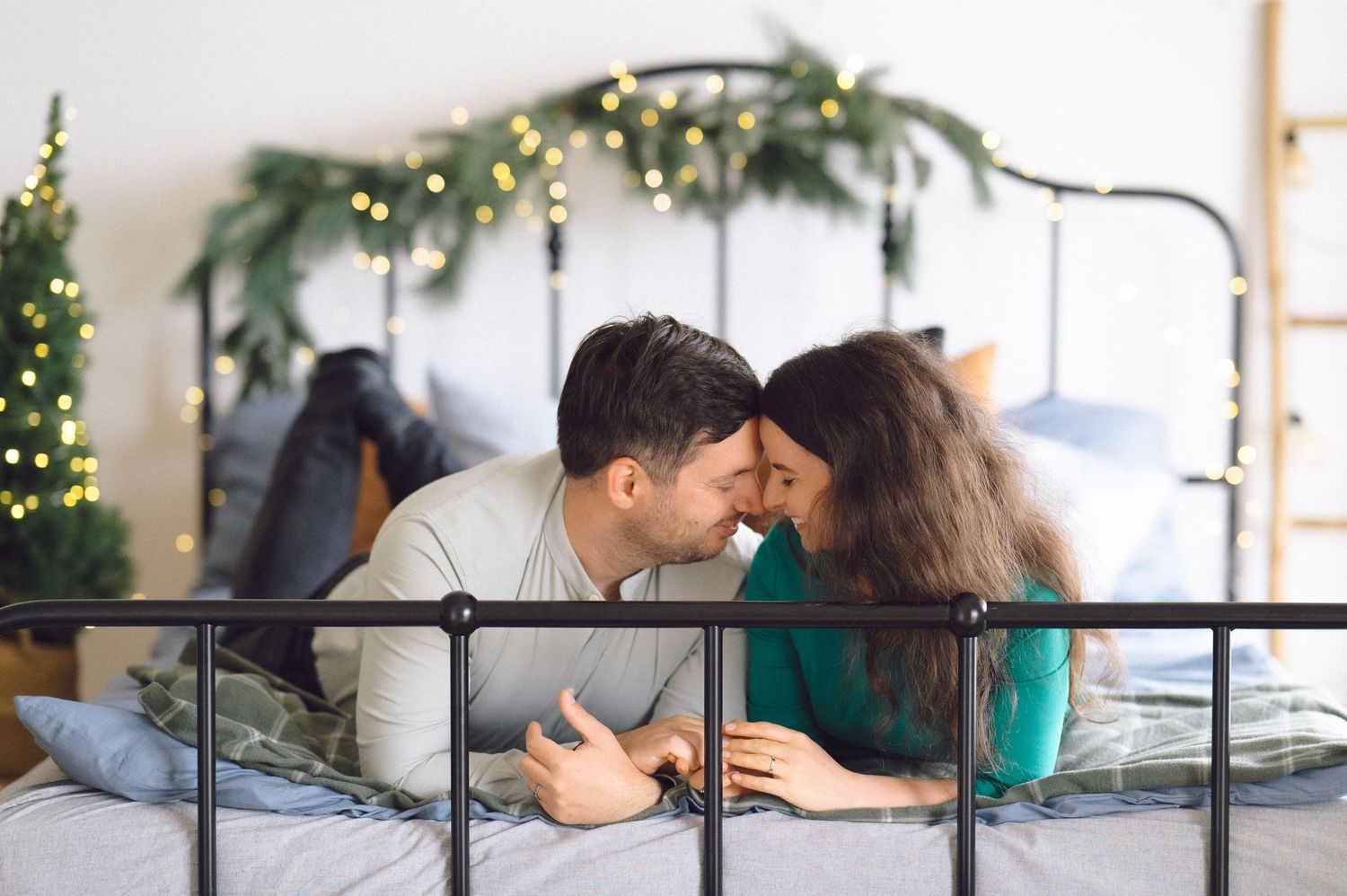 A romantic couple relaxes together on a bed with holiday decorations and greenery visible in the background.