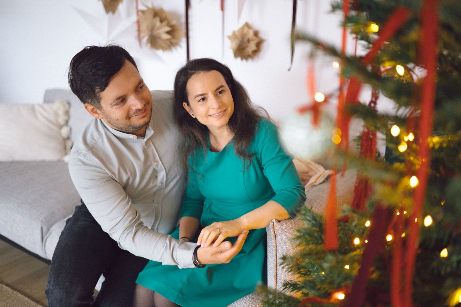 A couple in cozy winter attire share a tender moment near a decorated Christmas tree with twinkling lights in their home.