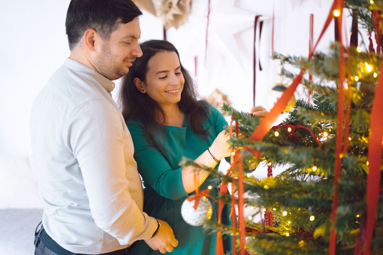 A couple in white and green decorates a Christmas tree together with ornaments and lights in a bright room.